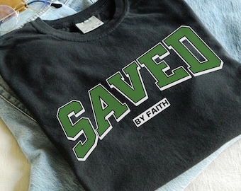 Puede incluir: Camiseta negra con la palabra "SAVED" en letras verdes grandes con contorno blanco. El texto "BY FAITH" está debajo en un rectángulo blanco. La camiseta está doblada sobre unos vaqueros azules.