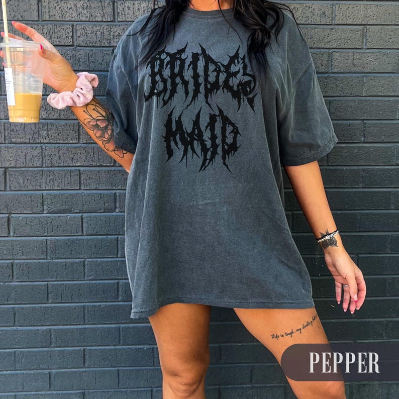 May include: A dark gray oversized t-shirt with the words "BRIDES MAID" in a gothic font. The shirt has short sleeves and a relaxed fit. The word "PEPPER" is in a banner at the bottom of the image.