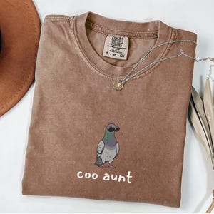 May include: A brown short-sleeved t-shirt with a crew neckline. The shirt features a graphic of a pigeon wearing black sunglasses and the text "coo aunt" printed in white below the bird. The interior neck label reads "Comfort Colors".