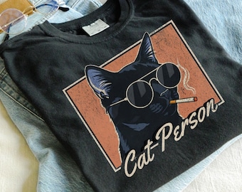 May include: Black t-shirt featuring a graphic of a black cat wearing sunglasses and smoking a cigarette, with the text "Cat Person" in a retro style. The shirt is folded on top of a pair of blue jeans and a pair of glasses.