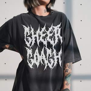 May include: Dark gray t-shirt with the white text "SHEER GANGSH" in a spiky, gothic font. The shirt has a vintage, worn look and a relaxed fit. The model is wearing the shirt.