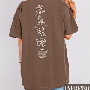 May include: Espresso-colored t-shirt with a vertical white graphic of beach-themed illustrations, including a seashell, surfboard, bikini, sand dollar, and tote bag. The word "ESPRESSO" is printed in a white rectangular box at the bottom.
