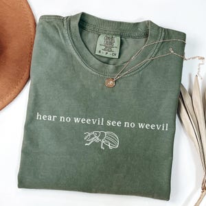 Könnte beinhalten: Ein olivgrünes T-Shirt mit dem Text "hear no weevil see no weevil" und einer Strichzeichnung eines Rüsselkäfers. Eine goldfarbene Halskette mit Anhänger und ein brauner Hut sind ebenfalls sichtbar.
