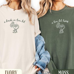 May include: Ivory and moss green t-shirts with a mermaid cocktail graphic. The ivory shirt has the text "a bride in tini bit", and the moss green shirt has the text "a tini bit tipsy". Both shirts have a relaxed fit.