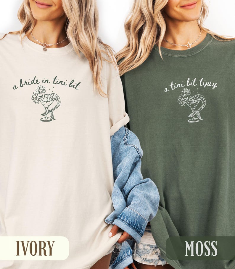 May include: Ivory and moss green t-shirts with a mermaid cocktail graphic. The ivory shirt has the text "a bride in tini bit", and the moss green shirt has the text "a tini bit tipsy". Both shirts have a relaxed fit.