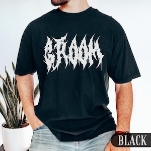 May include: Black t-shirt with the word "GROOM" in a white, spiky font. The shirt is a dark color and has short sleeves. The text is centered on the front of the shirt. The shirt is a casual style.