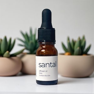 May include: A small amber glass bottle of santal diffuser oil with a black dropper. The label reads "santal diffuser oil 10mL Crafted in the USA." The bottle is set against a backdrop of potted succulents.