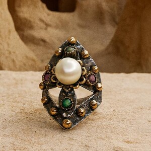 Puede incluir: Un anillo plateado con una perla blanca redonda y grande en el centro. El anillo presenta un diseño en forma de diamante con detalles dorados y pequeñas piedras preciosas de colores. El anillo está sobre una superficie marrón claro.
