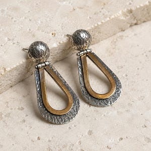 May include: A pair of drop earrings featuring a textured silver-tone spherical stud connected to a hinged teardrop-shaped pendant. The pendant consists of an outer silver-tone frame with a hammered finish and an inner smooth brass-tone teardrop loop.