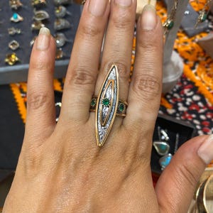 May include: A gold and silver ring with an elongated, leaf-like design. The ring features three emerald-green stones and intricate metalwork. Other jewellery pieces are visible in the background, including earrings and rings.