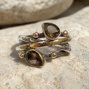 Op de afbeelding: Een ring met meerdere banden van geoxideerd zilver en goudkleurig metaal. Het ontwerp bevat twee peervormige, gefacetteerde bruine edelstenen en vier kleine, gladde goudkleurige bolletjes langs de overlappende, getextureerde banden.