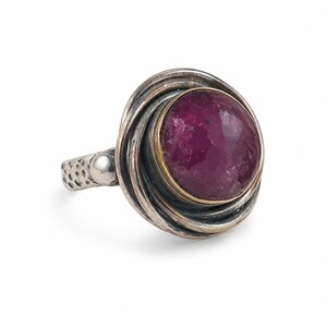 May include: A ring featuring a large, round, faceted deep magenta gemstone set in a gold-toned bezel. The stone is surrounded by multiple layers of coiled, oxidized silver wire. The band has a textured, hammered finish with a smooth silver bead.