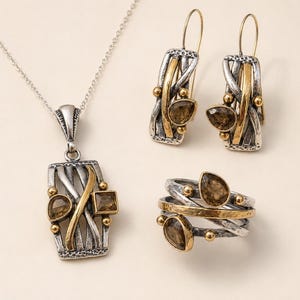 May include: A jewellery set consisting of a pendant necklace, a pair of drop earrings, and a ring. Each piece features a mixed-metal design with oxidised silver-tone textured bars, polished gold-tone accents, and faceted brown gemstone inlays.