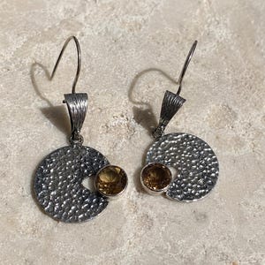 May include: A pair of silver-toned drop earrings featuring a circular, hammered metal disc with a textured surface. Each earring is accented with a single, round, faceted amber-colored gemstone set in a smooth bezel, suspended from a curved wire hook.