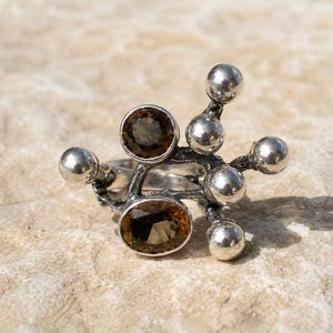 Op de afbeelding: Een zilveren ring met twee gefacetteerde, ovale bruine edelstenen in een textuurrijke, geoxideerde metalen zetting. Het ontwerp bevat diverse gladde, gepolijste zilveren bolletjes die uitsteken vanaf de centrale band, wat een asymmetrische, takachtige structuur vormt.
