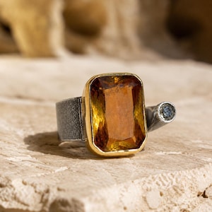 May include: A ring featuring a large, rectangular, amber-colored faceted gemstone set in a gold-toned bezel. The band is made of a textured, dark gray metal, and a small, round, light blue stone is set on a protruding arm of the band.