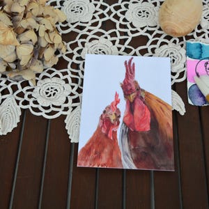 Può includere: Un dipinto ad acquerello di due galli con creste rosse e piume marroni e arancioni. L'opera d'arte è su una carta bianca, con un disegno rosa e blu ad acquerello e pennelli nelle vicinanze. L'immagine è su una superficie di legno scuro.