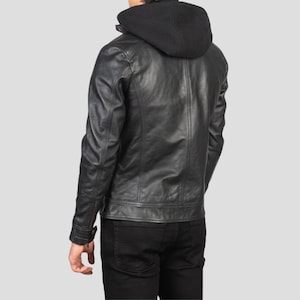 May include: A black leather jacket with a black hooded sweatshirt attached. The jacket has a classic design with a zipper closure and buttoned cuffs. The person is wearing black pants. The background is a solid light gray.