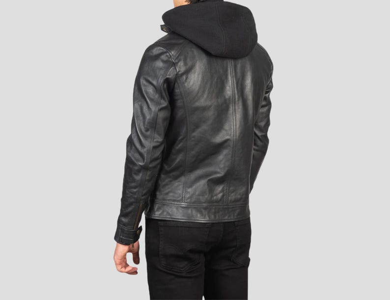May include: A black leather jacket with a black hooded sweatshirt attached. The jacket has a classic design with a zipper closure and buttoned cuffs. The person is wearing black pants. The background is a solid light gray.