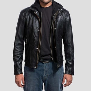 May include: Black leather jacket with a zip-up front, snap closures, and a hooded design. The jacket has a classic biker style with a tailored fit. The model is wearing blue jeans and a black shirt underneath.