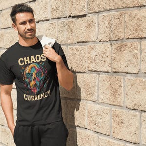 May include: Black t-shirt with the text "CHAOS CURRENCY" above and below a colorful skull graphic. The skull features blue, yellow, and red hues. The person is standing next to a beige brick wall.