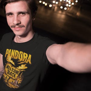 May include: Black t-shirt with the word "PANDORA" in gold lettering above a sign that reads "WELCOME BACK" in gold. The sign is set against a landscape with a sunset or sunrise. The shirt is worn by a person.