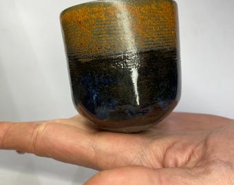 May include: A small, ceramic cup with a dark blue and orange glaze. The cup is hand-thrown and has a slightly uneven rim.