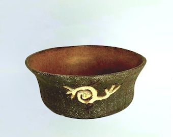 May include: A small, round, brown ceramic bowl with a white design of a stylized bird with a spiral on its body. The bowl has a rough, textured surface.
