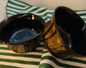 May include: Two ceramic bowls with a dark, glossy interior and a textured exterior. One bowl is upright, the other tilted, revealing a blue interior. The bowls rest on a green and white striped fabric.