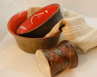 May include: Two brown ceramic bowls with red interiors, one nested inside the other, and a smaller brown ceramic cup with red streaks. The bowls are sitting on a striped beige and white cloth.