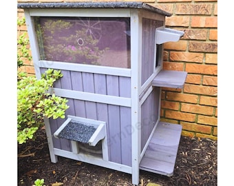 May include: A two-story wooden cat house painted in shades of gray and white. The structure features a window, a small roof, and two platforms. The cat house is placed next to a brick wall and surrounded by greenery.