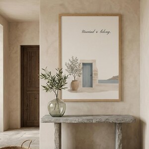 Può includere: Una stampa d'arte incorniciata che raffigura una scena mediterranea con una porta blu, un ulivo in vaso e il testo Unwind & belong. L'opera è esposta in una cornice di legno chiaro, sopra una console in pietra con un vaso di vetro.