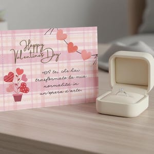 Può includere: Un biglietto di San Valentino con un motivo a quadri rosa e le parole "Happy Valentine's Day". Un piccolo portagioie aperto bianco sporco mostra un anello di diamanti. La carta ha anche un design a cuore.