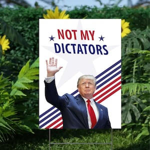 May include: A white yard sign with the text "NOT MY DICTATORS" and an image of a man in a suit. The sign has a metal stake for placement in a garden.