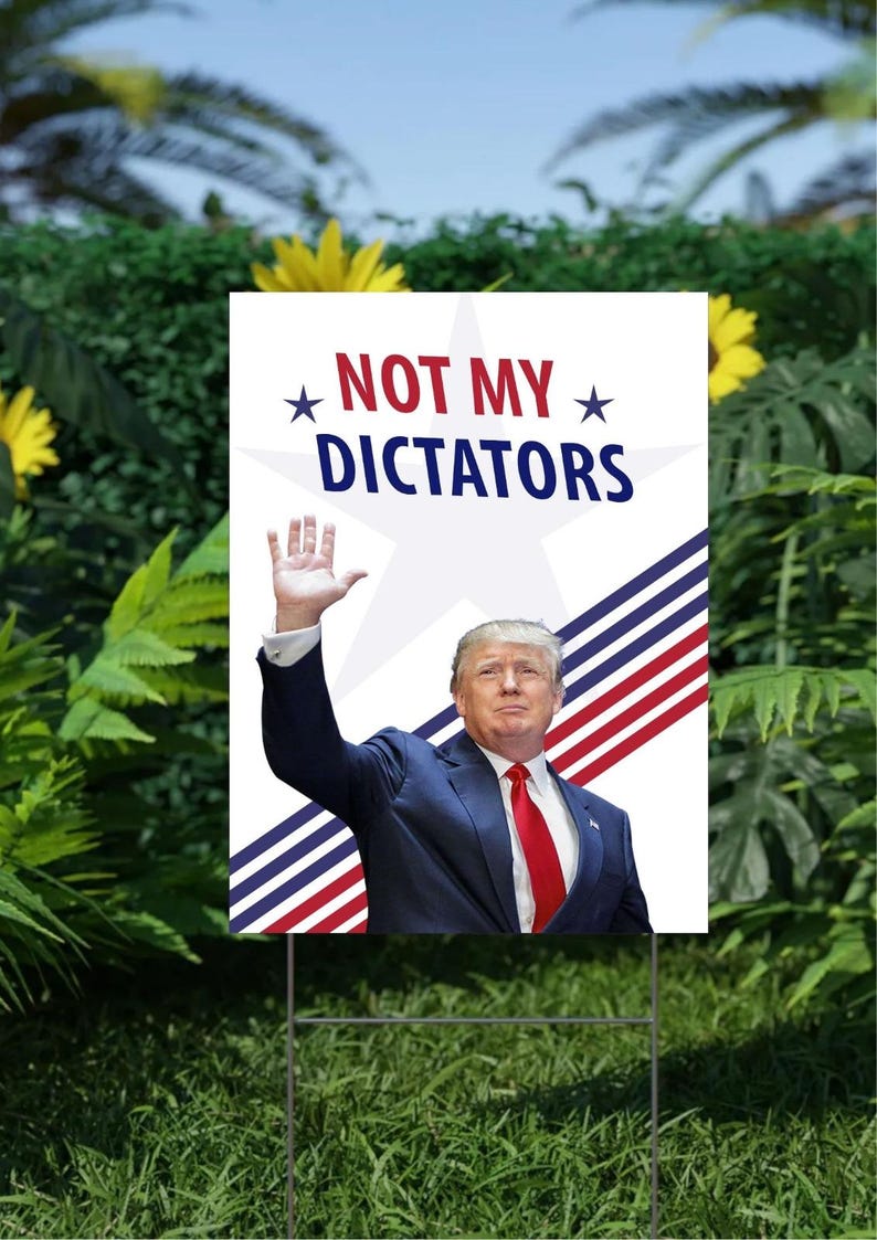 May include: A white yard sign with the text "NOT MY DICTATORS" and an image of a man in a suit. The sign has a metal stake for placement in a garden.