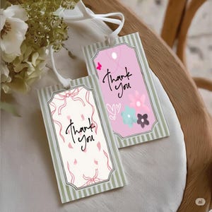 Op de afbeelding: Twee rechthoekige cadeaulabels met de woorden "Thank You" in cursief schrift. E&eacute;n label heeft een roze achtergrond met bloemen en een vlinder, de andere heeft een cr&egrave;mekleurige achtergrond met roze strikken en bloemblaadjes. Beide hebben groen-witte gestreepte randen.