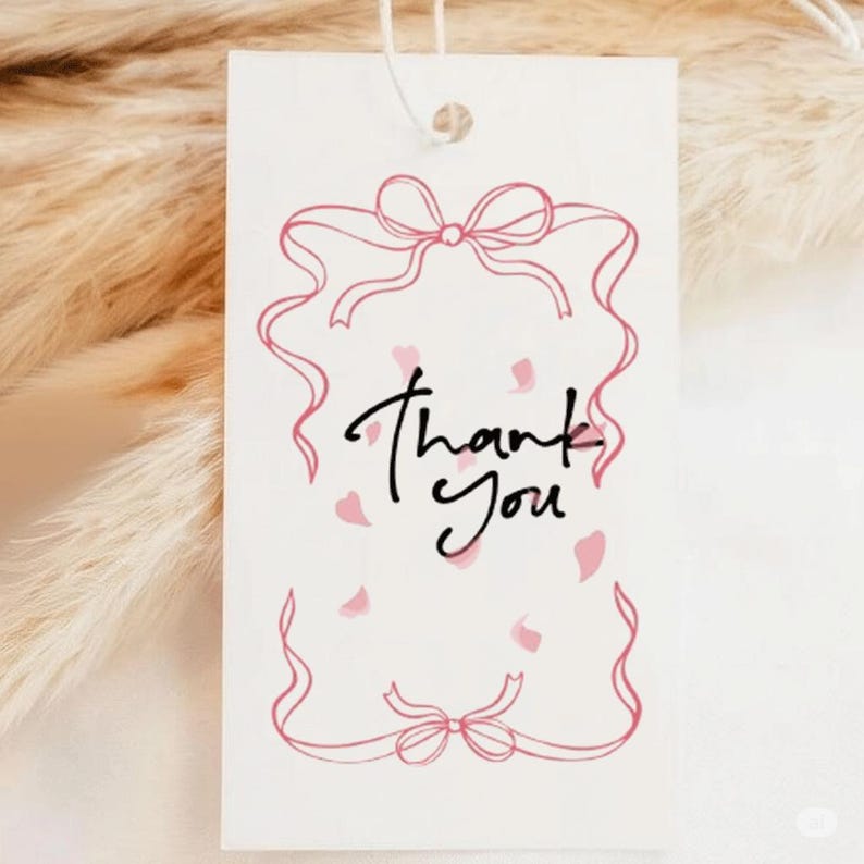 Op de afbeelding: Een wit cadeaulabel met een roze strik- en lintontwerp. De woorden "Thank You" zijn in zwart script geschreven. Roze bloemblaadjes zijn verspreid rond de tekst. Bovenop is een wit touwtje bevestigd.