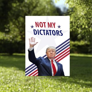 May include: A garden sign displaying the words "NOT MY DICTATORS" above an image of a man in a suit waving. The sign features a white background with red, white, and blue stripes and stars. It is set in a grassy garden.