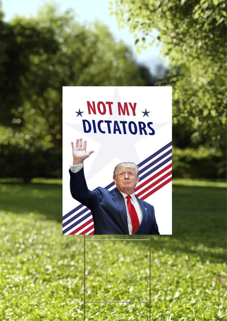 May include: A garden sign displaying the words "NOT MY DICTATORS" above an image of a man in a suit waving. The sign features a white background with red, white, and blue stripes and stars. It is set in a grassy garden.