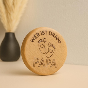 Può includere: Un disco rotondo di legno chiaro con le parole "WER IST DRAN?" e "PAPA" incise. Il disco presenta due contorni di piedini di bambino con dettagli a cuore. Un vaso nero con piante secche è sullo sfondo.