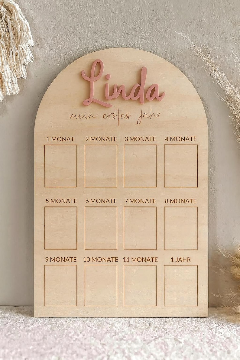 May include: Wooden milestone board with the name "Linda" in pink script. The board is arched at the top and has spaces for 12 months, labeled in German. The text "mein erstes jahr" is also present.