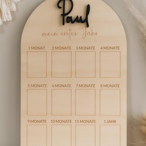May include: Wooden milestone board with the name "Paul" in black script. The board is arched at the top and has spaces for months 1-12, with the text "mein erstes Jahr" and month labels in German.