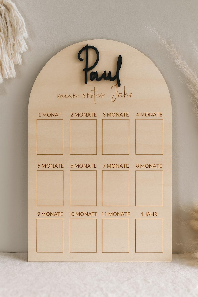 May include: Wooden milestone board with the name "Paul" in black script. The board is arched at the top and has spaces for months 1-12, with the text "mein erstes Jahr" and month labels in German.