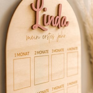May include: Wooden milestone board with the name "Linda" in pink script. The board has a light wood finish and features spaces for monthly milestones, with text in German. A decorative item for baby's first year.