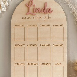 May include: Wooden milestone board with the name "Linda" in pink script. The board is arched at the top and has spaces for 12 months, labeled in German. The text "mein erstes jahr" is also present.