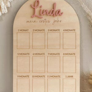 May include: Wooden milestone board with the name "Linda" in pink script. The board is arched at the top and has spaces for 12 months, labeled in German. The text "mein erstes jahr" is also present.