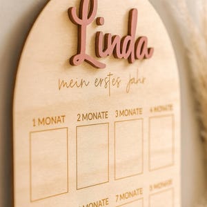 May include: Wooden milestone board with the name "Linda" in pink script. The board is light-colored with engraved text and boxes for tracking a baby's first year. The text "mein erstes jahr" is also engraved.