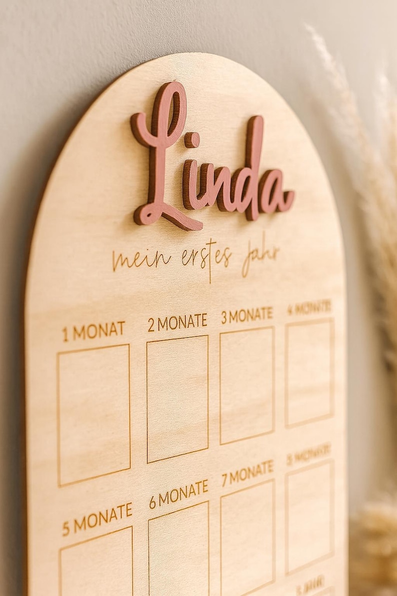 May include: Wooden milestone board with the name "Linda" in pink script. The board is light-colored with engraved text and boxes for tracking a baby's first year. The text "mein erstes jahr" is also engraved.