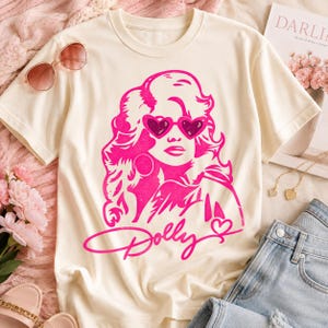 Könnte beinhalten: Ein cremefarbenes Kurzarm-T-Shirt mit einem leuchtend pinken Grafikdruck einer Person mit voluminösem Haar, die herzförmige Sonnenbrillen trägt. Unter der Grafik steht das Wort Dolly in Schreibschrift mit einem kleinen Herzsymbol.