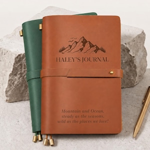 Puede incluir: Un diario de cuero color canela con un gráfico de montaña, el texto "HALEY'S JOURNAL" y una cita grabada en la portada. Un diario de cuero verde se encuentra detrás y un bolígrafo de metal dorado descansa sobre la superficie junto a los diarios.